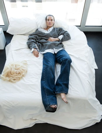 Model Charlotte Tomas lies on a white bed in a grey knit sweater and blue jeans, with a blonde wig beside her.