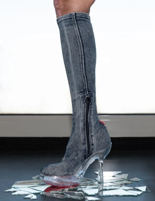 Fashion-forward denim knee-high stiletto boot stepping on shattered glass, set against a gradient white-to-gray backdrop, creating a dramatic scene.
