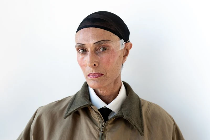 Close-up studio portrait of an adult woman with a black net cap, tan jacket, white collared shirt, and dark tie against a white backdrop.