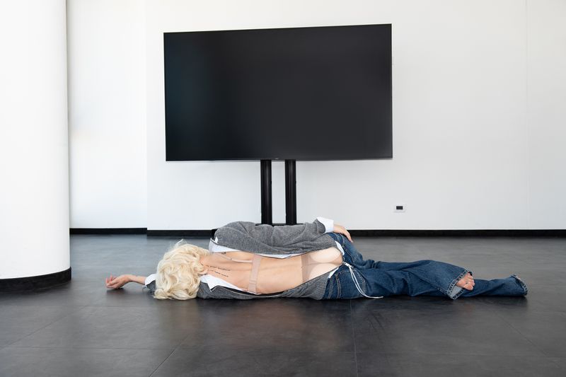 An adult female sprawls on the floor in front of a large flat-screen TV. She is wearing jeans, a grey sweatshirt, and tan hose.