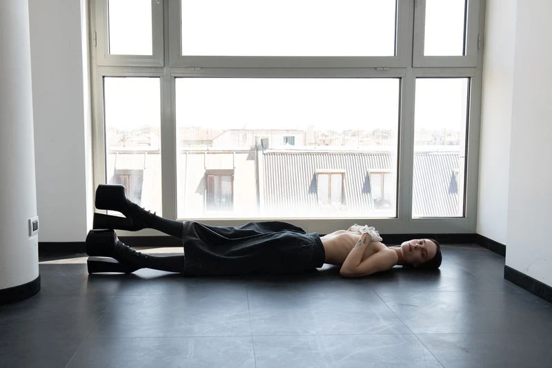 A young adult female lies on a black floor near a window wearing a black skirt and chunky platform boots in an indoor environment.