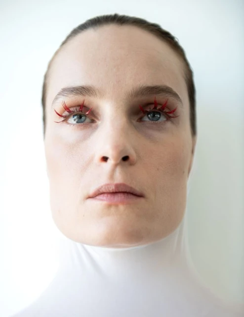 Close-up studio portrait of a young fair-skinned woman with bright blue eyes wearing spiky red false eyelashes in a beauty context.