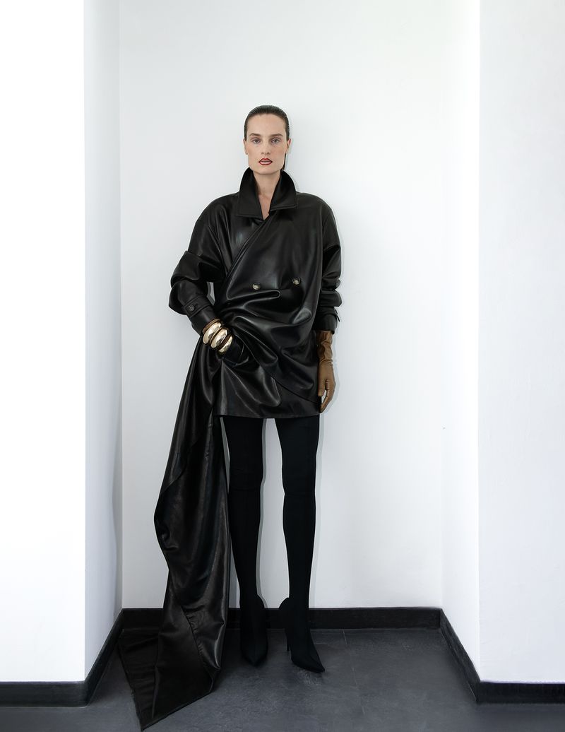 Fashion portrait of a woman in a black leather coat and black leggings posing in a white corner with gold jewelry on her wrists.