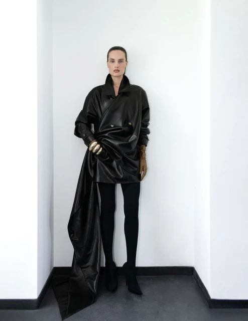 Fashion portrait of a woman in a black leather coat and black leggings posing in a white corner with gold jewelry on her wrists.