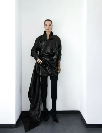 Fashion portrait of a woman in a black leather coat and black leggings posing in a white corner with gold jewelry on her wrists.