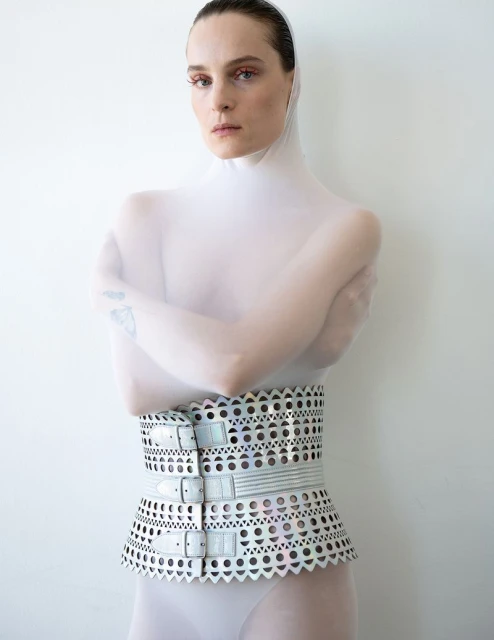 A fair-skinned adult female model is posing indoors wearing a semi-sheer full bodysuit and futuristic metallic belt with buckles against a white background.
