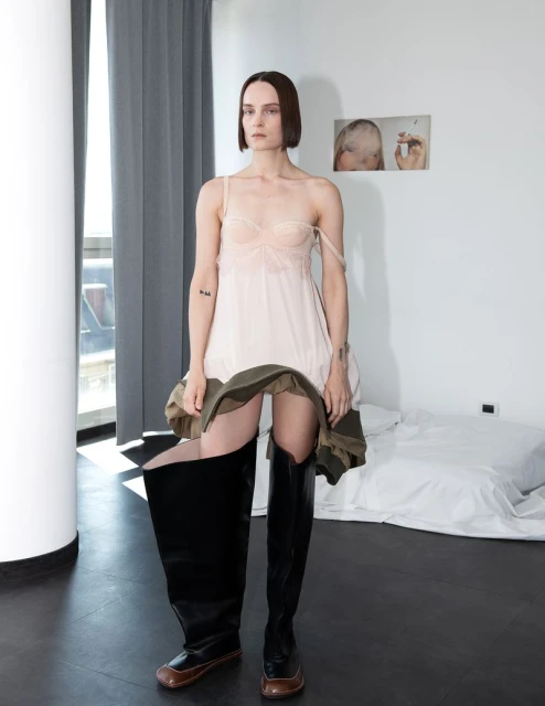 Adult female fashion model poses in a light pink slip dress and large black boots inside an apartment with a bed and large windows.
