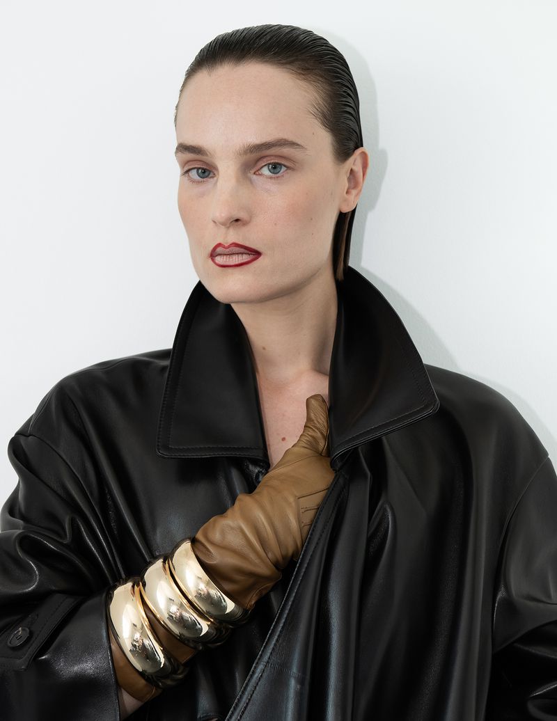 Fashion portrait of an adult woman with slicked-back hair wearing a black coat, gold bracelets, and tan gloves in a studio.
