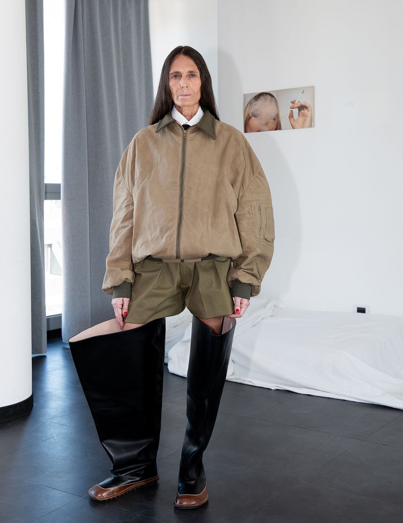 Full studio shot of a mature woman in an oversized tan jacket and thigh-high black boots posing in a minimalist studio apartment.
