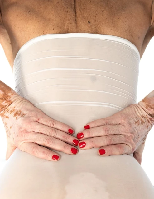 Back view of a woman wearing a waist shaper, with hands on her lower back, red nail polish, and a white background in a close-up shot.