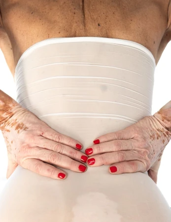 Back view of a woman wearing a waist shaper, with hands on her lower back, red nail polish, and a white background in a close-up shot.