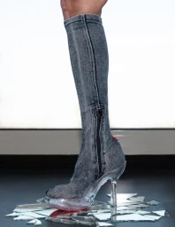 Denim high-heel knee boot with a clear acrylic platform stepping on shattered mirror glass on a dark studio floor.