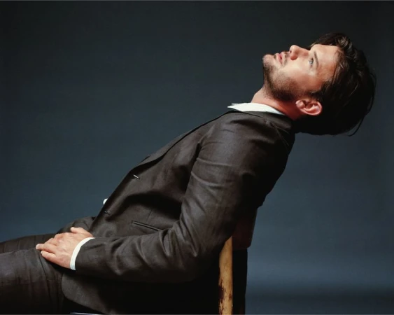 A man in a suit sits on a chair, leaning back with his head tilted up and to the side against a grey background.