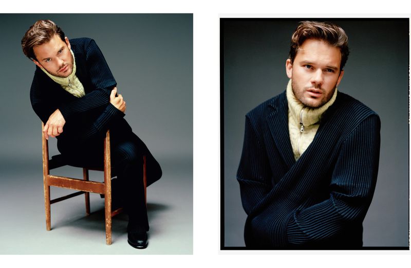Indoor portrait of a young adult male in a dark pinstripe suit and light-yellow turtleneck, seated on a wooden chair and posing for camera.