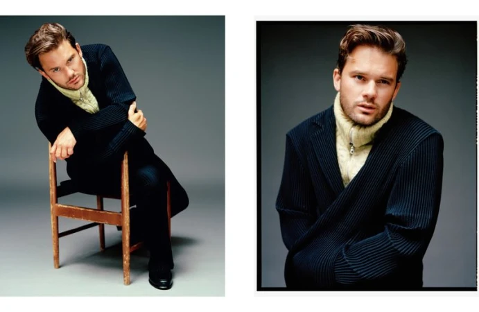 Indoor portrait of a young adult male in a dark pinstripe suit and light-yellow turtleneck, seated on a wooden chair and posing for camera.