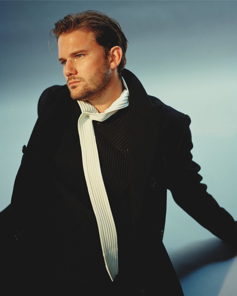 Portrait of a fair-skinned adult male in a dark coat and textured shirt, looking off to the side against a gradient blue background.