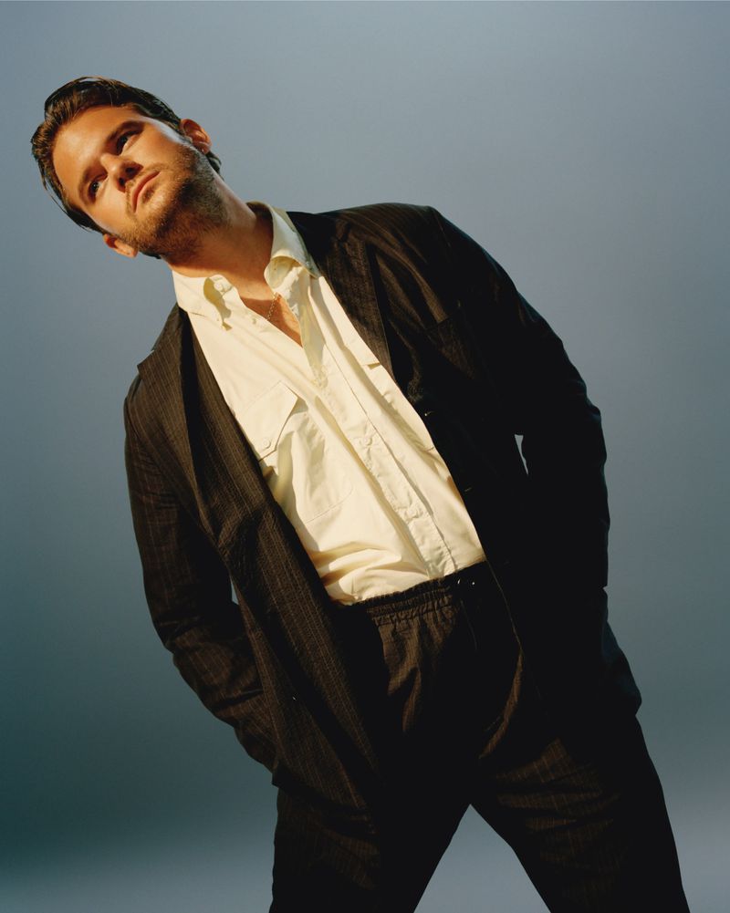 Portrait of a man in a black pinstripe suit and white dress shirt against a gradient blue background in a studio environment.