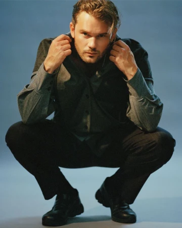 Jeremy Irvine crouching in a dark shirt and trousers against a grey studio seamless backdrop, holding his collar.