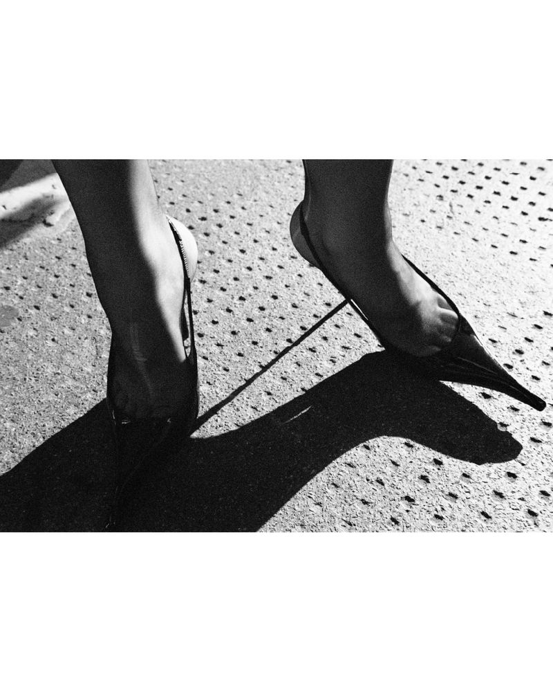 A pair of feet wearing slingback heels on a concrete surface, with a strong light source creating dark, defined shadows.