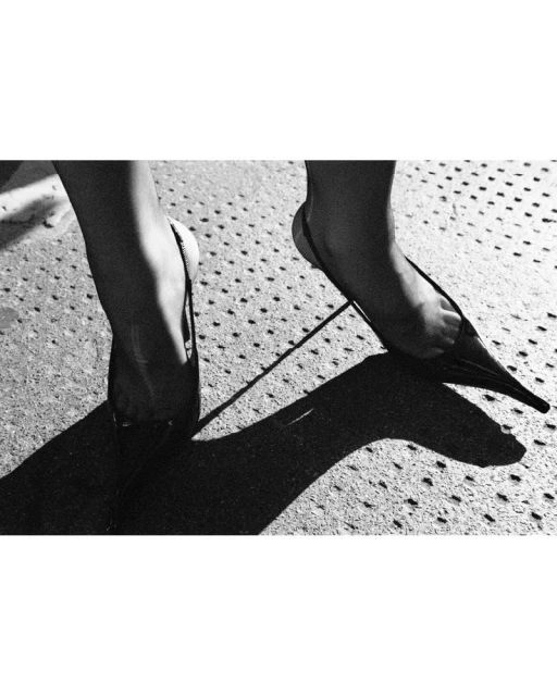A pair of feet wearing slingback heels on a concrete surface, with a strong light source creating dark, defined shadows.