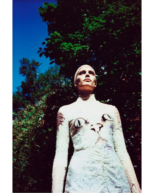 Adult female fashion model in an animal-print dress with cat face design posing outside against trees and a blue sky in a fashion editorial.