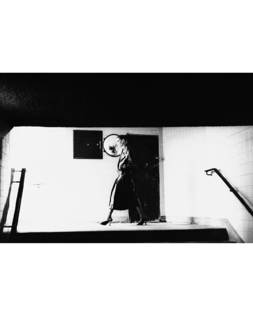 Monochromatic shot of an adult female in an overcoat and heels walking in a stark, indoor stairwell with minimalist architectural details.