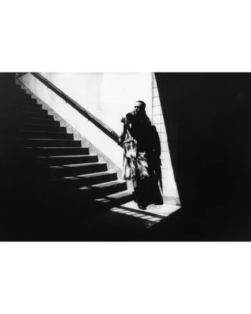 Monochrome image of a person in a long coat walking down stairs with dramatic lighting and shadows on the steps and walls.
