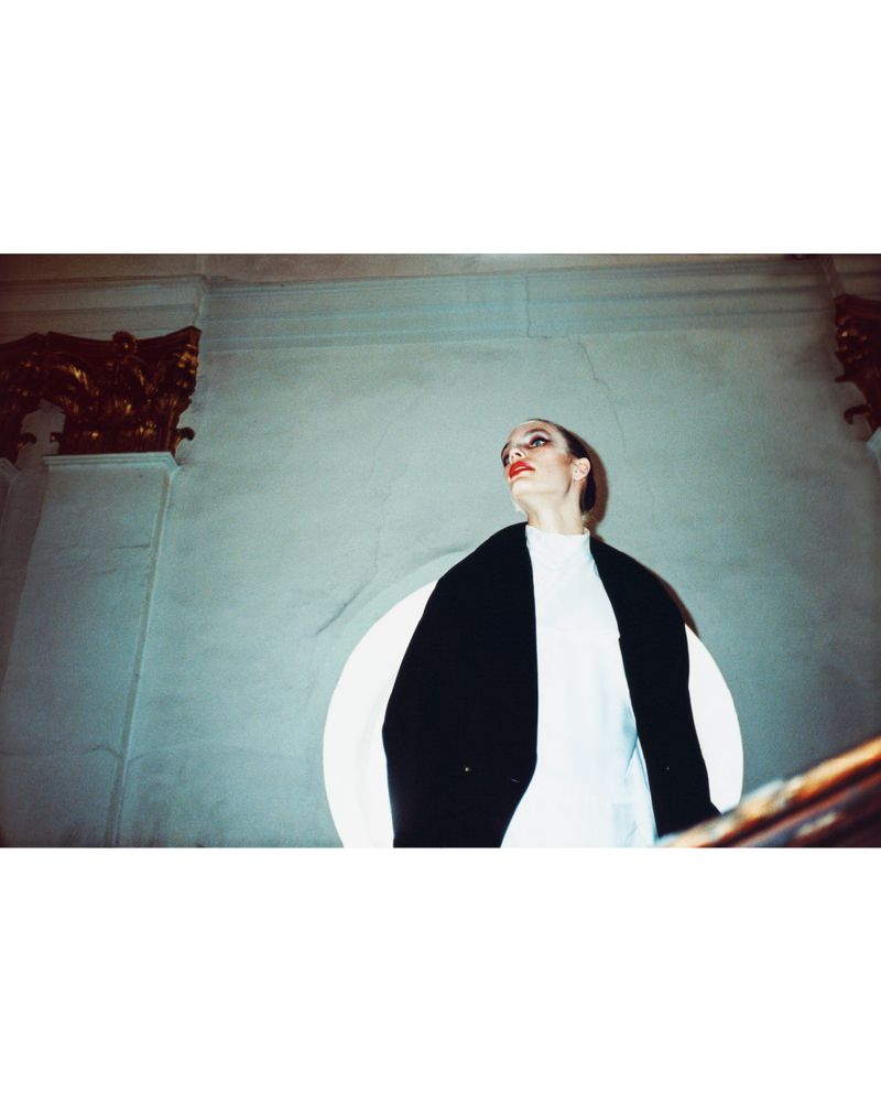 A woman in a white turtleneck and oversized black coat stands in a stairwell with gold-painted architectural details, looking upwards.