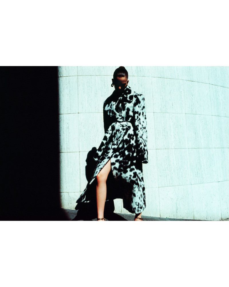 Model Betsy Gaghan wears a black and white mottled coat posing against a curved tiled wall in high-contrast light.