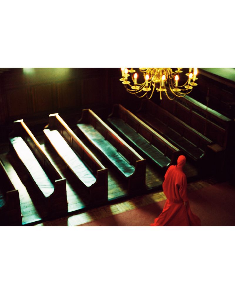 Interior view of church with wooden pews and a chandelier, a person in a red robe is walking in the aisle of the church.