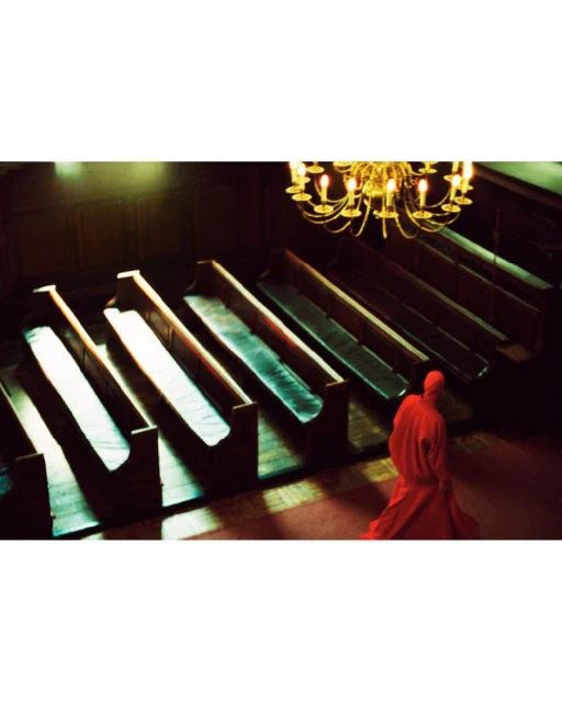 Interior view of church with wooden pews and a chandelier, a person in a red robe is walking in the aisle of the church.