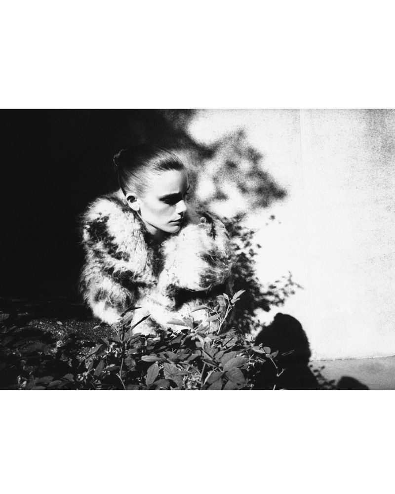 Black and white portrait of a model wearing a patterned textured coat outdoors amongst foliage, photographed by Chris Rhodes.