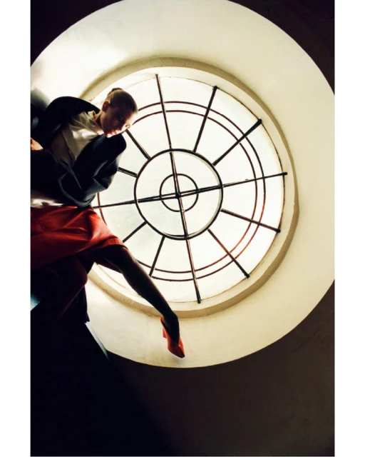 Fashion shot from low angle of woman in red dress and jacket, posing by round window. Stylish clothing and unique perspective.