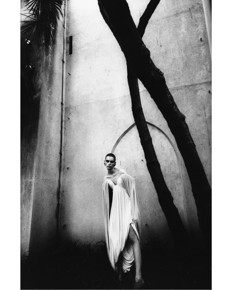 Model Betsy Gaghan in a draped white gown standing beneath large tree branches against a high architectural wall.