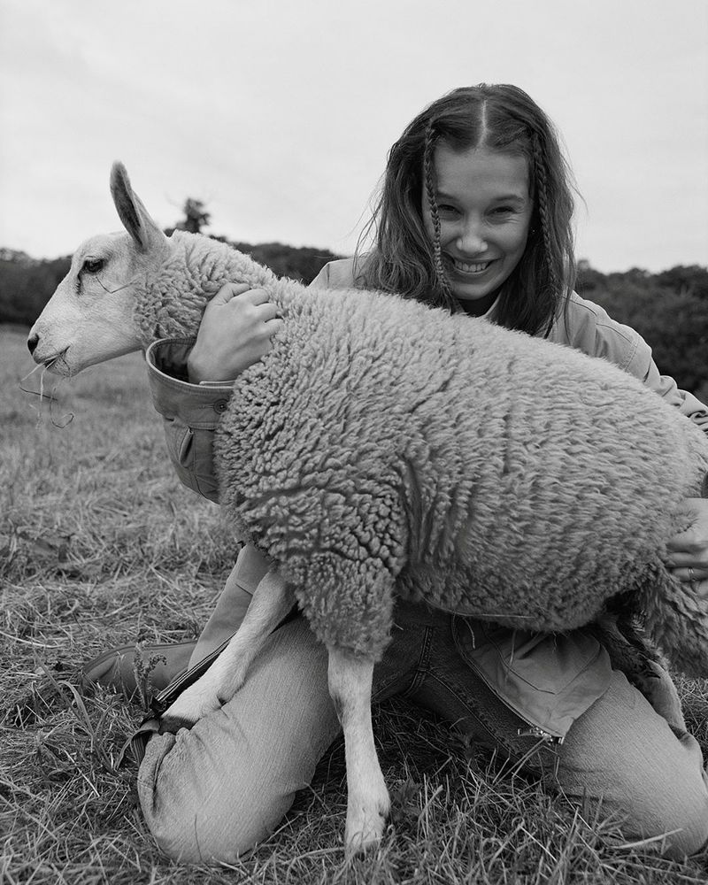 Young woman smiles while kneeling in a field and holding a sheep in her arms, set against a backdrop of cloudy sky and trees.