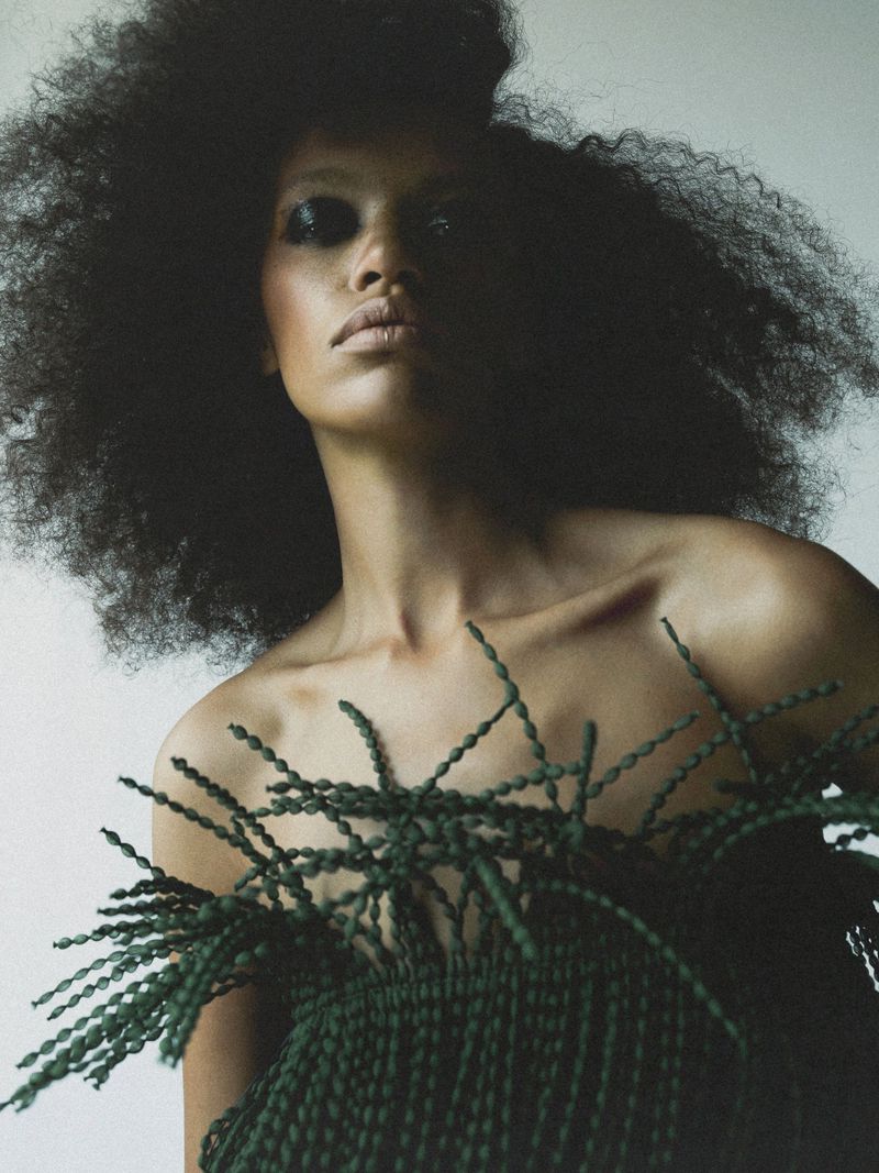 Portrait of a woman with dark hair in a large afro, posing in front of a neutral background wearing a rope top.