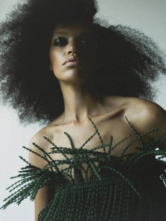 Portrait of a woman with dark hair in a large afro, posing in front of a neutral background wearing a rope top.