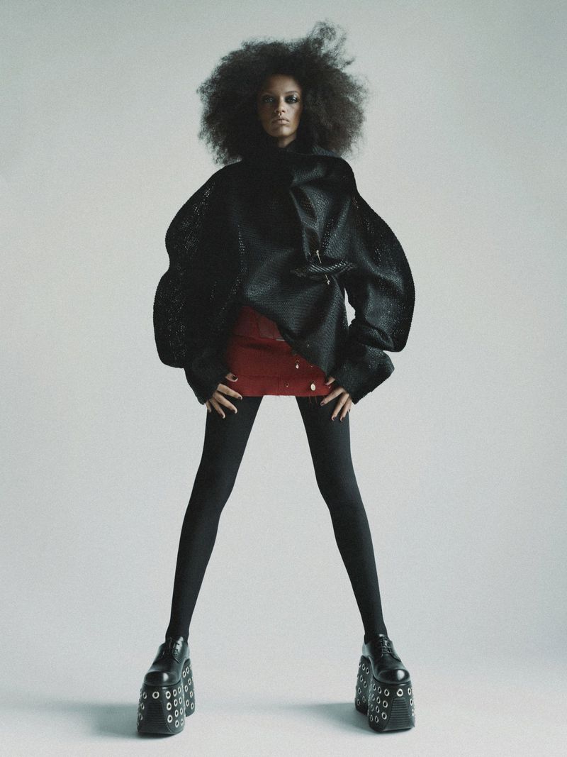 Fashion shot of a Black adult female in avant-garde clothing featuring a large afro, jacket, mini skirt, tights, and platform shoes. 