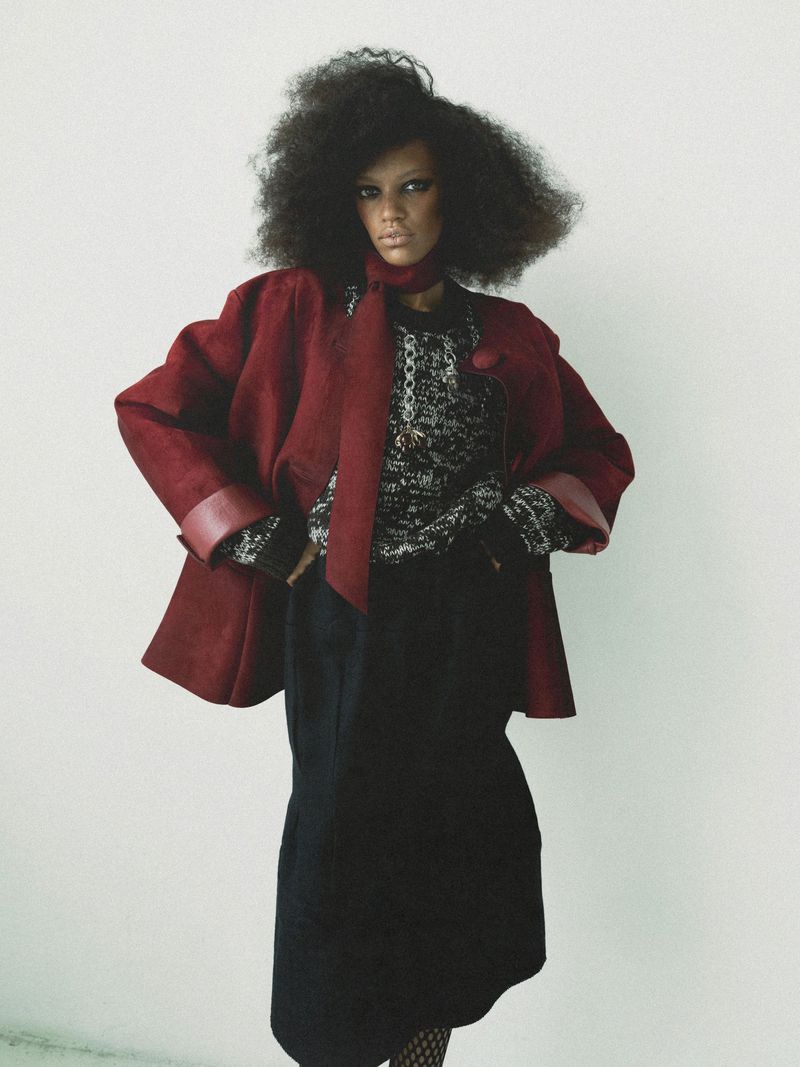 Fashionable adult woman in a studio wearing a maroon jacket and black skirt posing against a white background in a full shot.