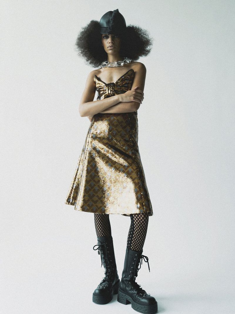 Elegant Black woman poses in a gold sequin dress, wearing platform boots and a distinctive hat against a minimalist white backdrop.