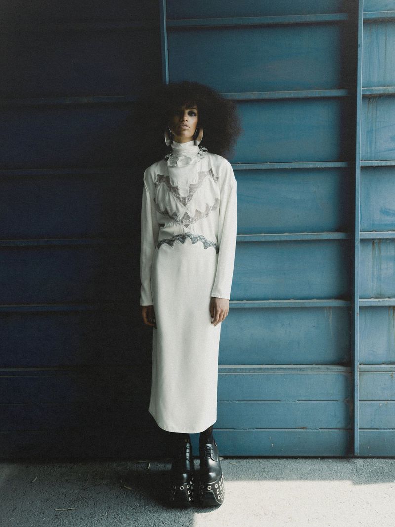 Kukua Williams in a high-neck white midi dress and platform boots posing against a blue slatted metal wall.