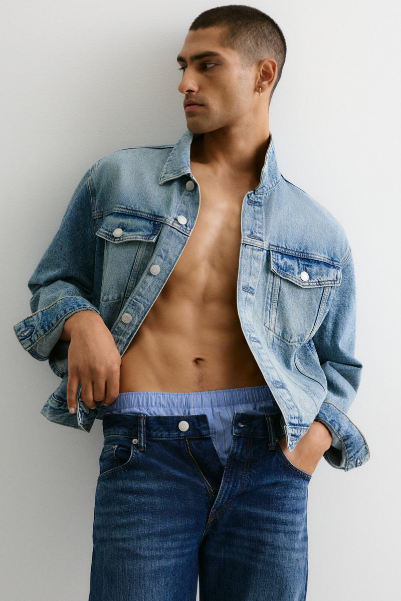Man poses in denim jacket and jeans against a white wall, showcasing fashion apparel in a studio setting with natural lighting.
