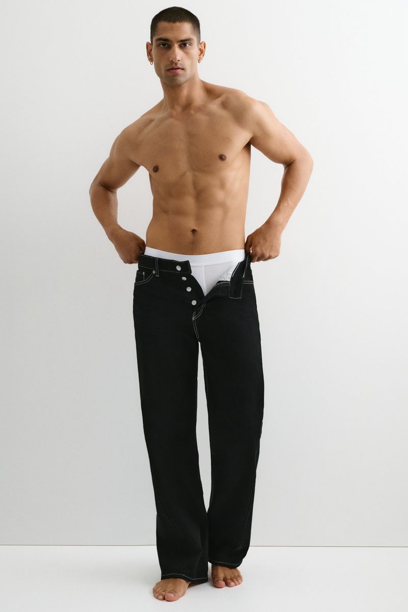 Shirtless man with tanned skin stands indoors, wearing black jeans and white underwear, posing in front of a bright white background.