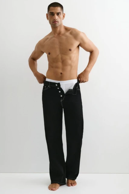 Shirtless man with tanned skin stands indoors, wearing black jeans and white underwear, posing in front of a bright white background.