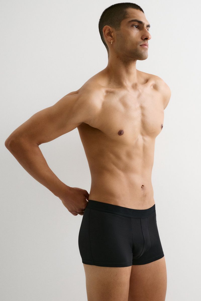 Athletic man with bare torso poses indoors with hand at waistband wearing black underwear against a plain white background. 