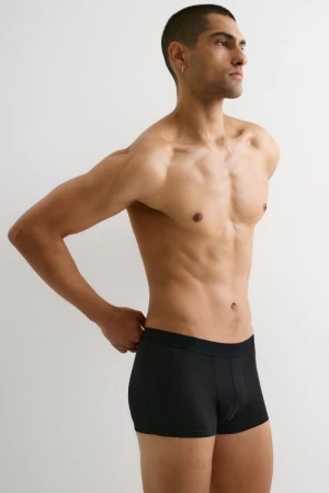 Athletic man with bare torso poses indoors with hand at waistband wearing black underwear against a plain white background. 