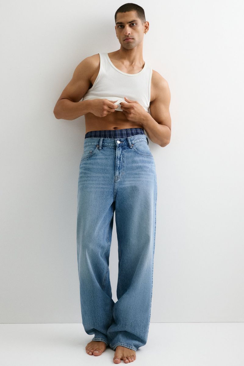 Adult male wearing a white tank top and light-wash denim baggy jeans stands barefoot against a white wall in a studio setting.