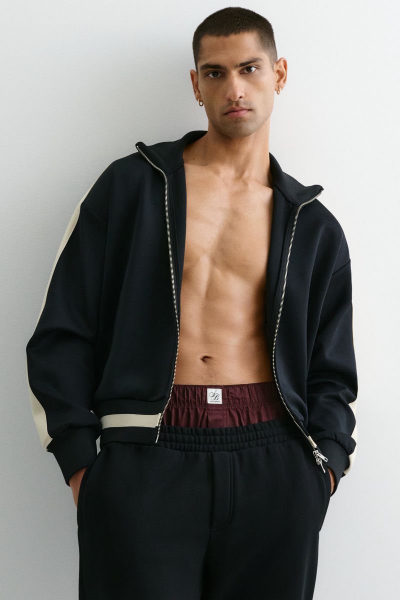 A man in a zip-up jacket and sweatpants stands indoors against a white wall, showcasing an athletic casual style with minimalist accessories.