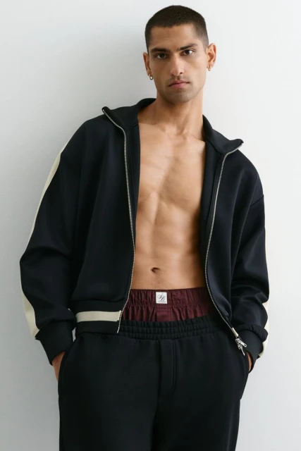 A man in a zip-up jacket and sweatpants stands indoors against a white wall, showcasing an athletic casual style with minimalist accessories.