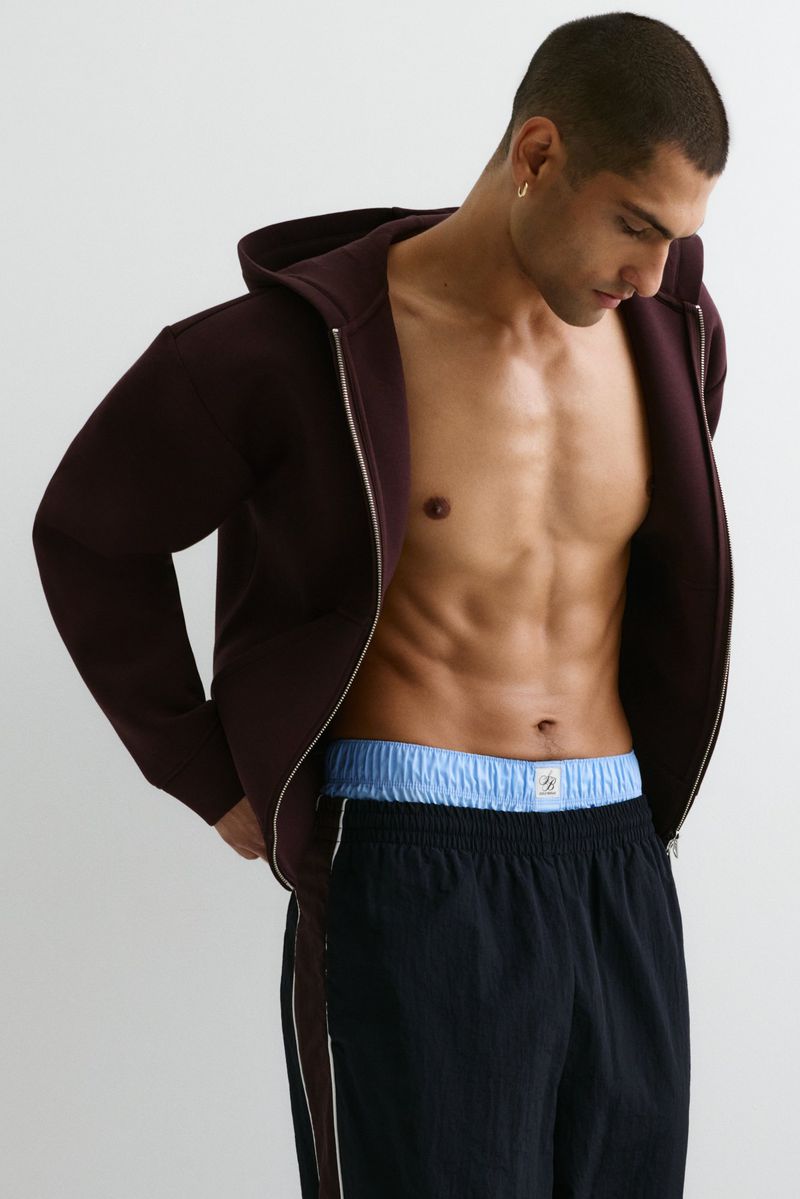 Adult male posing in an open plum-colored zip-up hoodie with black shorts against a light background in a medium shot.
